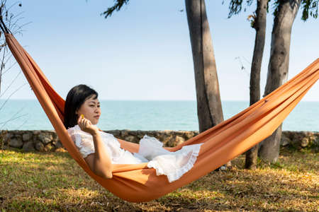 Summer vacations concept, Happy asian woman with white dress, sleeping rest and relaxing in hammock on tropical beach at sunshine dayの写真素材