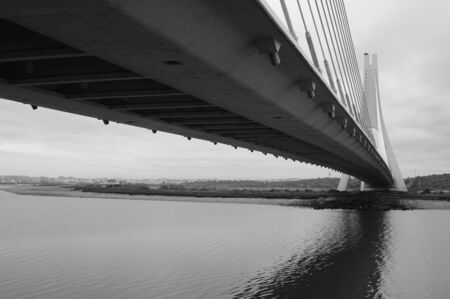 Black and white suspension bridge. Bank in the background and the brdge reflection can be seen in the waterの写真素材