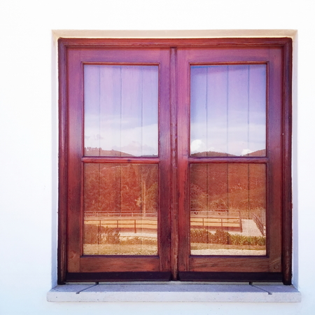 Square closed wood window on bright white wall with background reflected in the glass
