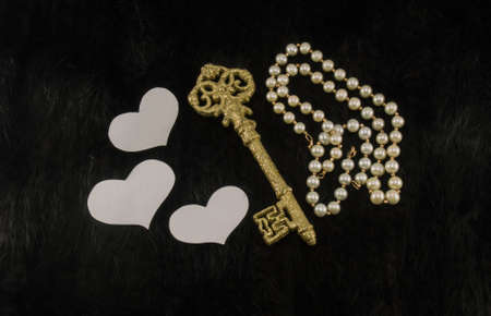 Old golden key with white paper hearts and pearl necklace on a black fur background