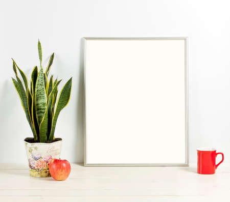 Silver frame mockup with plant pot, mug and apple on wooden shelf. Empty frame mock up for presentation design. Template framing for modern art.の写真素材
