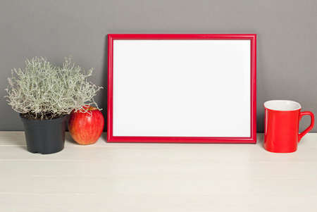 Red frame mockup with plant pot, mug and apple on wooden shelf. Empty frame mock up for presentation...