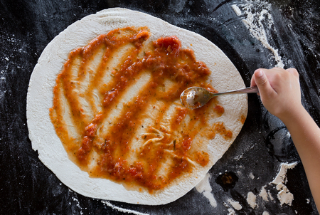 The kid spread the sauce on pizza dough on black tableの写真素材