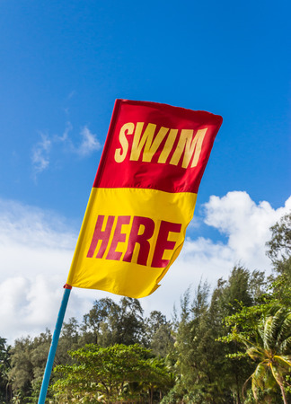 Red and yellow Swim Here safety flag with blue sky on the beach in Phuket, Thailandの写真素材