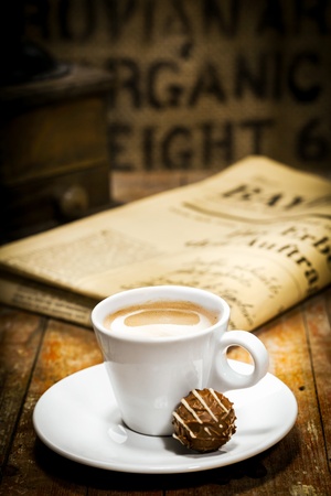 Cup of rich milky coffee with a chocolate bonbon on the saucer and a folded newspaper on the table behind in a coffee break conceptの写真素材