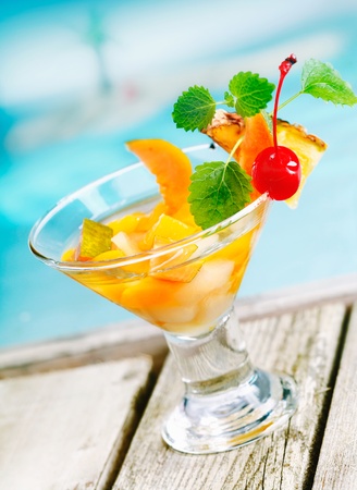 Refreshing colourful tropical fruit cocktail appetiser in a conical glass filled with pieces of fresh mixed fruit on decking alongside a blue poolの写真素材
