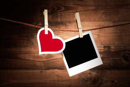 A red and white heart hangs from a wooden clothespeg alongside a blank instant photo frame on a rustic wooden background for your nostalgic romantic memories or Valentines messageの写真素材