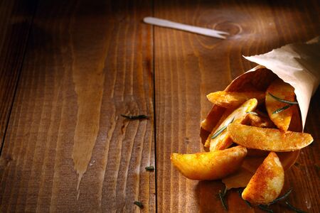 Tasty spicy potato wedges with fresh rosemary leaves spilling out of a rolled paper takeaway container onto textured wooden boards with copyspaceの写真素材