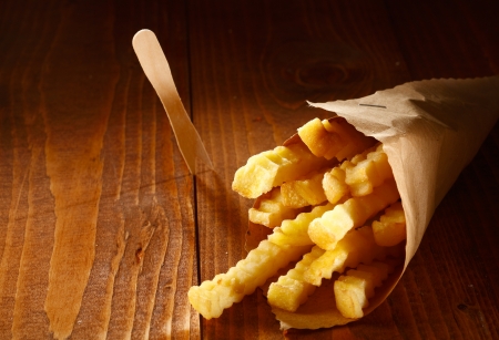 Crinkle cut golden french fries served as a takeaway in a roll of brown paper lying on a wooden...