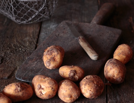 Fresh unwashed potatoes on a rustic chopping board with an old kitchen knifeの写真素材