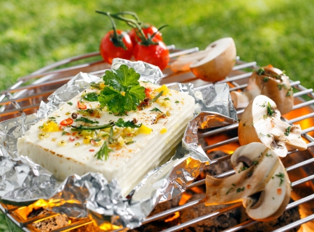 Large portion of seasoned halloumi or feta cheese grilling in tin foil over hot coals in a barbecue outdoors on green grassの写真素材