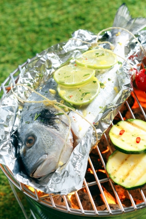 Whole fresh gilthead breem garnished with sliced lemon roasting on aluminium foil over the glowing coals in a barbecueの写真素材