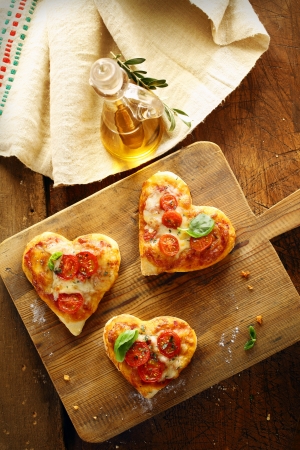 Cute heart shaped mini pizzas topped with cheese and tomato and garnished with fresh basil served on an old wooden board in the kitchen with virgin olive oilの写真素材