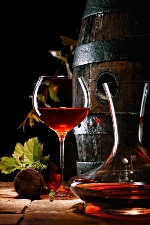 A glass of red wine next to a decanter with an oak cask in the background.の写真素材