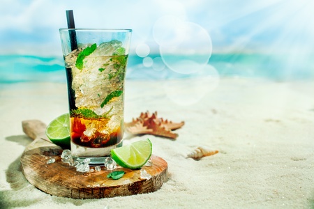 Refreshing iced rum, lime and mint mojito cocktail standing in a tall glass on an old wooden board on the golden sand of an idyllic tropical beach to celebrate a summer vacationの写真素材