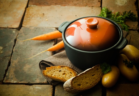 Closed casserole dish with fresh carrots and potatoes and slices of crusty bread on weathered bricks with copyspaceの写真素材