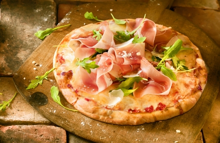 Preparing a proscuitto and rocket pizza with thinly sliced slices of ham and fresh rocket leaves arranged on a cheese and tomato paste base on a wooden paddle ina pizzeriaの写真素材