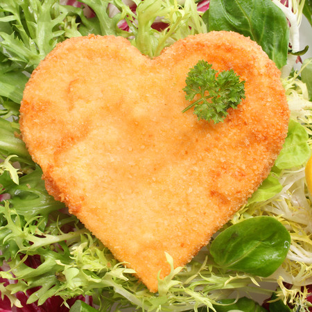 Romantic heart shaped fried golden schnitzel in breadcrumbs served on a bed of fresh leafy green lettuce and basil and garnished with parsleyの写真素材