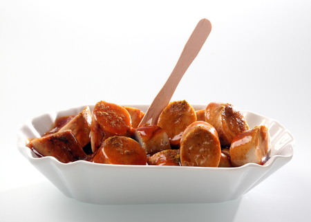 Appetizer of smoked sliced sausage drenched in a savoury sauce and served with a small wooden fork in a white fluted dishの写真素材