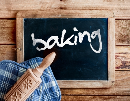 Country kitchen with a decorative carved wooden rolling pin with a floral pattern on oven gloves lying on an old vintage school slate with a distressed wooden frame with the handwritten word - bakingの写真素材