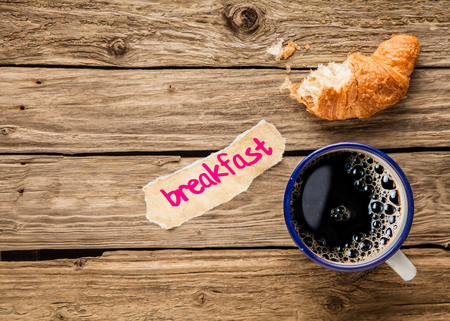 Breakfast - a handwritten note on a small fragment of torn paper lying alongside an aromatic cup of frothy full roast espresso coffee and a three quarter eaten freshly baked flaky croissantの写真素材