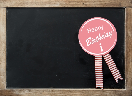 Happy Birthday rosette with festive striped ribbons on a vintage kids school slate with a wooden frame and blank black surface with copyspace for your textの写真素材