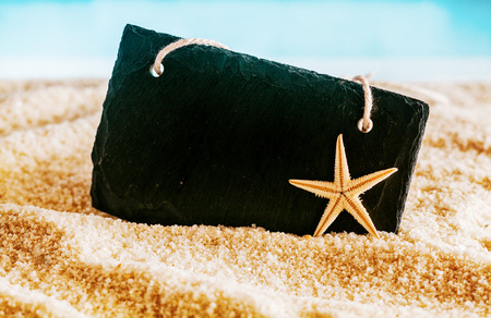Blank black sign with copyspace for your text or advertisement nestling on golden sea sand on a tropical beach in summer with a starfish and ocean backdropの写真素材