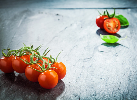 Fresh grape tomatoes on the vine in the lower corner of a dark textured wooden surface with highlight and vignetting and a halved tomato in the top corner with copyspace betweenの写真素材