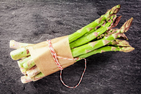 Bundle of farm fresh green asparagus spears tied with brown paper and string on a dark textured slate background, overhead viewの写真素材