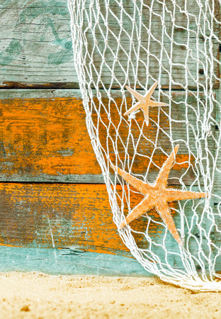 Rustic marine background with starfish suspended in a fishing net over weathered blue and orange painted wooden boards with copyspace and beach sand belowの写真素材