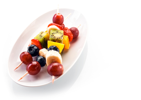 Fresh healthy tropical fruit kebabs with grapes, blueberries, banana, orange, kiwifruit and strawberries served on a white oval dish on a white background with copyspace, high angleの写真素材