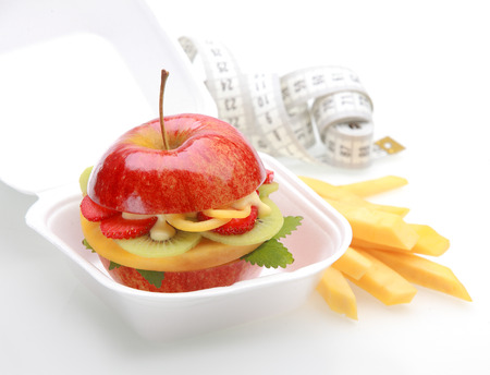 Healthy apple burger takeaway in a fast food container with a juicy red apple sliced through and filled with diced fresh tropical fruit and sauce with fruit batons or chips and a tape measureの写真素材