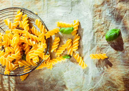 Preparing an Italian fusilli pasta dish with fresh basil leaves and the traditional spiral corkscrew shaped pasta lying in an old retro wire strainer on grunge crumpled paper outdoors in the sunshineの写真素材