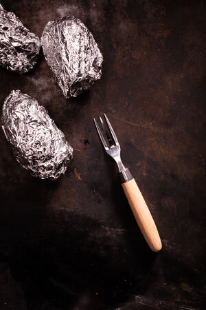 Barbecued foil wrapped potatoes still wrapped in their silver aluminium foil laying on an old slate surface with a fork and copyspace, overhead viewの写真素材