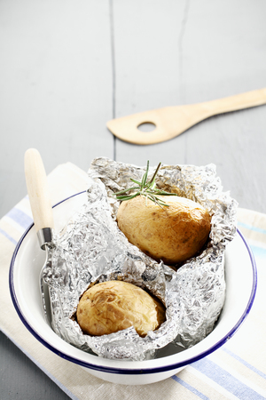 Fresh hot baked potatoes with rosemary still nestling in their foil wrappers in a metal basin on a white painted wooden picnic table, with copyspaceの写真素材