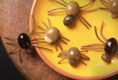 Cute spooky Halloween spider appetizers made from green, black and stuffed olives with spaghetti legs crawling off a yellow platter onto a wooden table, view from aboveの写真素材