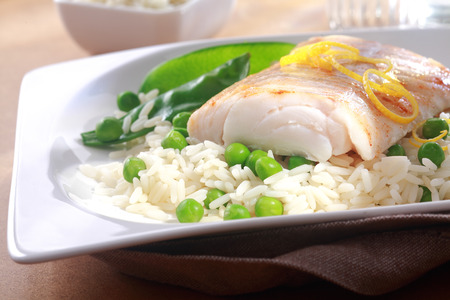 Healthy meal of baked fish fillet garnished with fresh lemon zest, rice and peas served on a white plateの写真素材