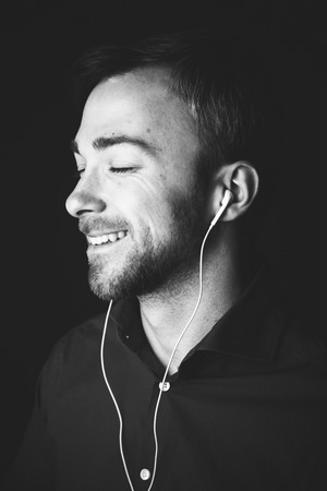 Close up head and shoulders greyscale portrait of a handsome cute charismatic guy listening to music on a set of earplugs with a smile of bliss and pleasureの写真素材