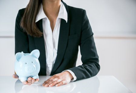 Conceptual Businesswoman in Black and White Suit, Sitting at the Table with a Sky Blue Piggy Bank on her Hand, Captured Close up.の写真素材