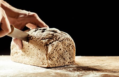 Man cutting a freshly baked crusty homemade loaf of wholegrain bread with a knife on a wooden...