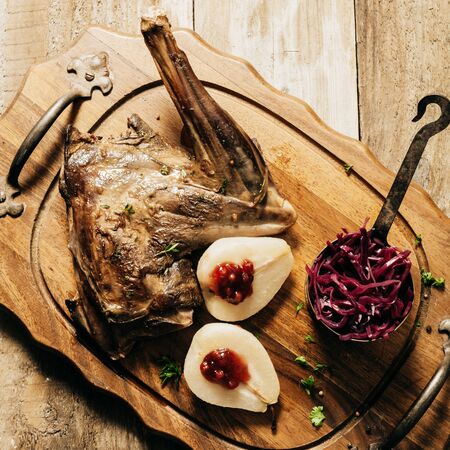High Angle View of Roasted Venison Haunch on Wooden Tray with Prepared Pears on Rustic Tableの写真素材