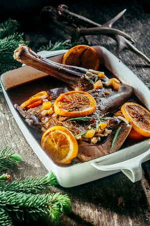 Roasted Venison Haunch in Roasting Pan Seasoned with Fresh Herbs and Orange Citrus Slices on Rustic Wooden Surface Surrounded by Evergreen Sprigs and Deer Antlersの写真素材