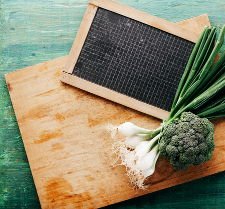 High Angle View of Fresh Onions and Broccoli on Wooden Cutting Board with Black Board Marked with Grid Lines with Rustic Green Backgroundの写真素材