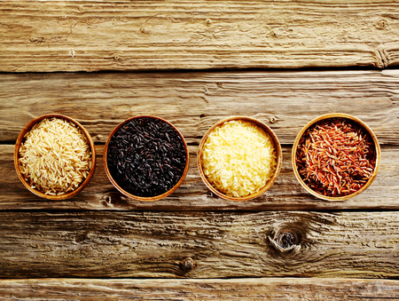 Four different varieties of dried rice, wild, basmati, long-grain and red, displayed in individual wooden bowls in a line on an old rustic weathered wooden table, overhead view with copyspaceの写真素材