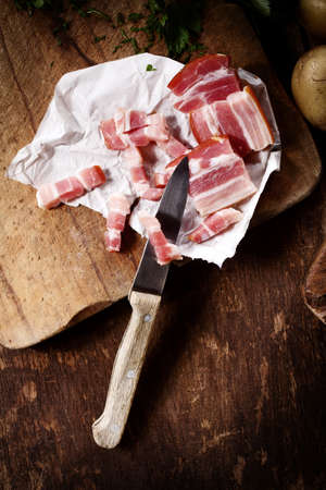 Dicing cured smoky bacon on a wooden cutting board for use in savory cooking on an old rustic wooden table top in the kitchen, view from above with a knifeの写真素材