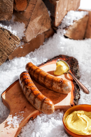 Grilled sausages served on a rustic wooden board with a scoop of mustard outdoors in the snow at a winter grill partyの写真素材