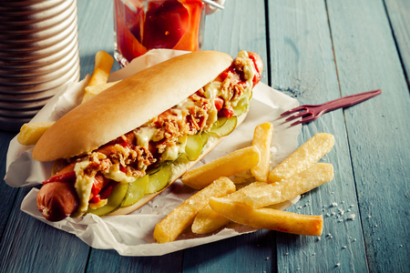 Tasty hot dog with a smoked frankfurter sausage covered with full trimmings on a bed of cucumber on a fresh roll with golden crispy fried potato chips and relish or ketchup on a rustic wooden tableの写真素材