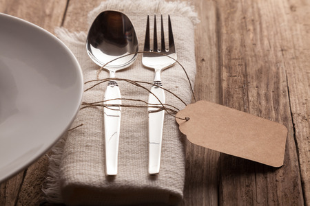 Close Up of Silver Spoon and Fork Tied with Twine and Blank Tag on Natural Napkin with Fringed Edges Beside Neutral Plate and Set on Rustic Wooden Table with Copy Spaceの写真素材