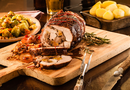 Small pork roast cut and sliced with visible stuffing on cutting board next to dishes of potatoes and brussels sprouts