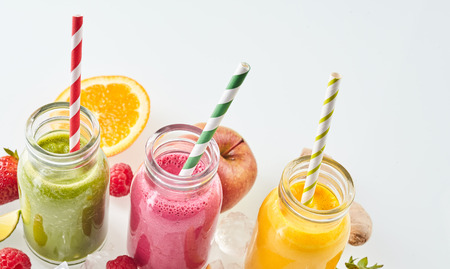 Angled row of apple, mango and strawberry smoothie beverages surrounded by ice cubes and various pieces of fruit over gray background with copy spaceの写真素材